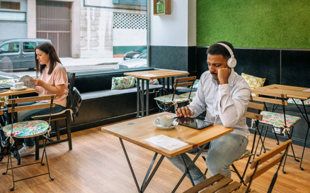 Freelancers working at various tables, engaging with their devices and sipping coffee in a modern cafe featuring large windows and a vibrant green wall, embodying urban life and creativityの写真素材