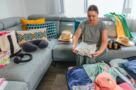 Happy woman packing clothes into a suitcase while sitting on a sofa in her living room, eagerly preparing for an exciting summer vacation travel adventureの写真素材