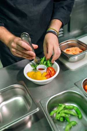Close up of unrecognizable chef hands placing ingredients with tongs in a delicious and healthy poke bowl, like salmon, edamame, tomatoes, mango and sauce, working meticulously in restaurant kitchen.の写真素材