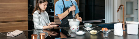 Banner of two teenagers are making a cake in modern kitchen, one using hand mixer while other consults a recipe on tablet. Brother and sister cooking together. Family leisure time at home concept.の写真素材