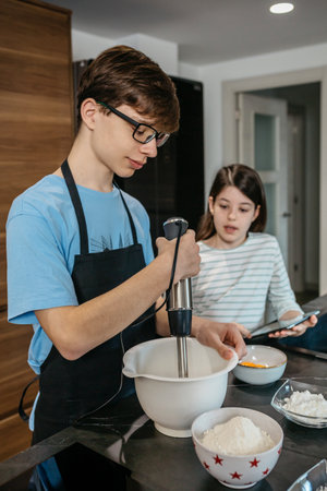 Teenage boy using hand blender while mixing ingredients in bowl, following recipe on tablet held by sister in kitchen, enjoying family moment and learning culinary skills. Happy time cooking concept.の写真素材