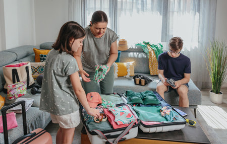 Family folding and packing clothes and beachwear into suitcases while teenage male using digital camera, getting ready for summer vacation. Mother and siblings preparing bags for trip in living room.の写真素材