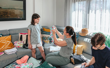 Happy mother showing colorful t shirt to her smiling daughter while folding and packing clothes into suitcases, getting ready for summer vacation. Family preparing bags for a trip in living room.の写真素材