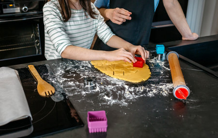 Unrecognizable young girl cutting cookies from dough with various cookie cutters while boy helping to her in a modern kitchen. Brother and sister making festive dessert together for a party at homeの写真素材
