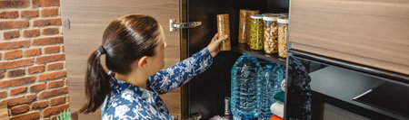 Banner of woman preparing emergency food pantry in kitchen for disasters. Female placing cookies package to 72 hours survival kit stored in cupboard. Alert, foresight and self sufficiency strategy.の写真素材
