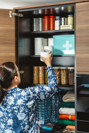 Unrecognizable female filling toilet paper to stocked emergency food pantry in kitchen, preparing for potential disasters. 72 hours survival kit concept. Alert, foresight and self sufficiency strategyの写真素材