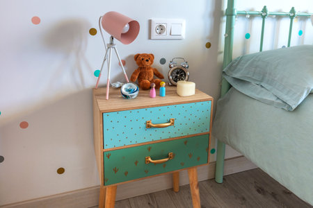 Modern childs nightstand featuring a teddy bear, a pink lamp, an alarm clock, decorative figurines and a heart-shaped candle, adding a touch of warmth and personality to the bedroomの写真素材