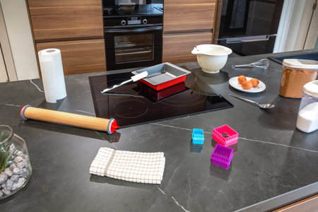 Baking ingredients like eggs and sugars, along with utensils such as a rolling pin and cookie cutters, are neatly arranged on a modern kitchen counter with an induction cooktopの写真素材
