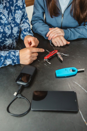 Unrecognizable mother pointing at multi tool knife while explaining 72 hours emergency kit to teenage daughter with power bank, lighter and mobile. Alert, foresight and self sufficiency strategy.の写真素材