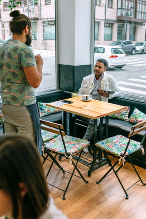 Waiter taking an order from customer using a tablet in a cozy cafe, featuring floral chairs and a wooden table, located near a window with a pleasant street viewの写真素材