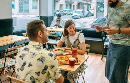 Happy couple talking and ordering food to waiter in restaurant while are sitting at table with pizza, salad and drinks and another customer is sitting in the background.の写真素材