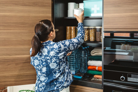 Unrecognizable female preparing 72 hours survival kit for potential disasters. Woman placing toilet paper to stocked emergency food pantry in kitchen. Alert, foresight and self sufficiency strategy.の写真素材