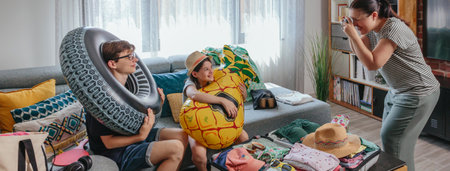 Banner of mother taking photo of her smiling children holding inflatable floats, with open suitcase full of clothes, swimwear and accessories. Happy family preparing luggage for summer vacation.の写真素材
