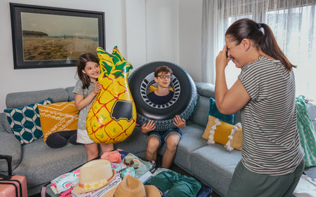 Smiling children holding inflatable floats and mother taking a photo next to suitcase full of clothes. Family having fun while packing luggage for vacation, in happiness and excitement environment.の写真素材