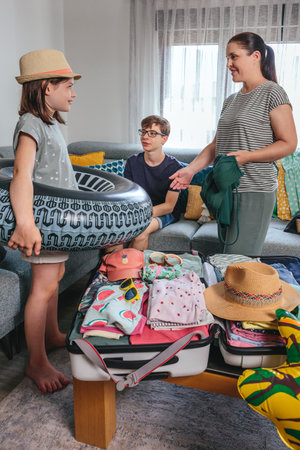 Smiling girl holding an inflatable giant tire float and mother with swimwear in her hand preparing suitcase for holidays. Mother, son and daughter packing luggage for summer vacation in living room.の写真素材