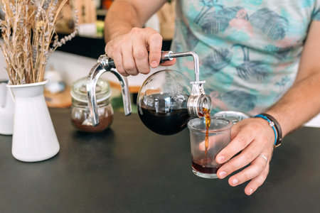 Close up of unrecognizable barista expertly pours brewed coffee from Japanese siphon coffee maker into a glass, showcasing the art of specialty coffee preparation.の写真素材