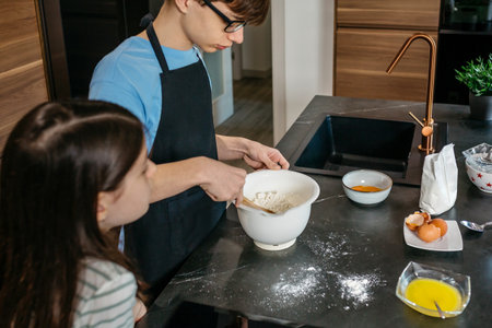 Boy and young girl preparing together homemade cake, mixing ingredients in bowl and sharing culinary learning experience with joy and teamwork. Brother and sister baking cake in modern kitchen at homeの写真素材