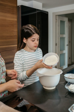 Concentrated teenage girl pours flour into a mixing bowl while baking in a modern kitchen surrounded by various ingredients. Young female child learning cooking skills at home concept.の写真素材