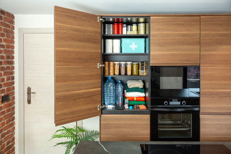 Survival pantry stored according to government recommendations about emergency or war preparedness. Cupboard with canned food, water bottles, first aid supplies and clothes in modern kitchen.の写真素材