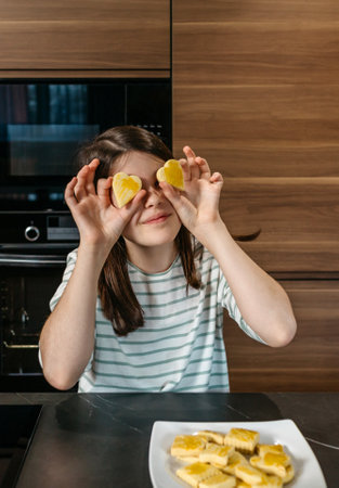 Young girl having fun in the kitchen, holding two heart shaped cookies over her eyes, expressing playfulness and enjoyment. Happy little girl looking at camera while covering her eyes with biscuits.の写真素材