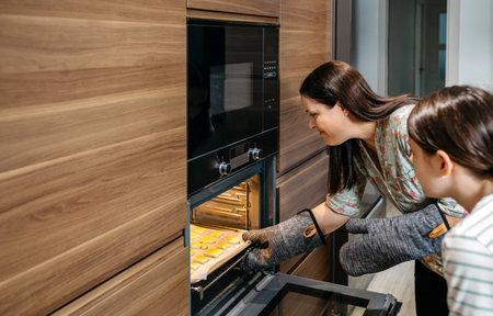 Mom and her daughter are taking a baking pan full of golden cookies out of the oven in their kitchen, wearing oven mitts and working together to carefully remove the freshly baked treatsの写真素材