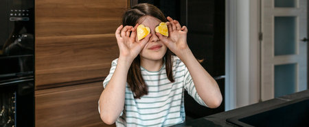 Banner of young girl having fun in the kitchen, holding heart shaped cookies over her eyes, expressing playfulness and enjoyment. Happy little girl looking at camera while covering eyes with biscuits.の写真素材