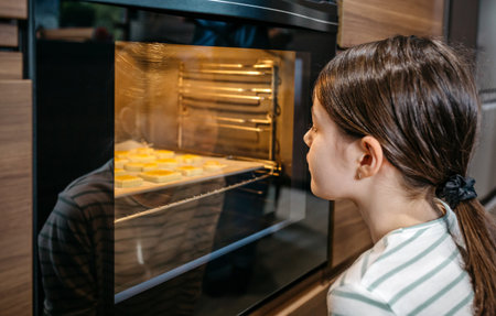 Little girl watching cookies baking in oven, waiting for them to be ready, enjoying the sweet smell and anticipating the delicious taste. Cooking at leisure time concept. Left copy space available.の写真素材