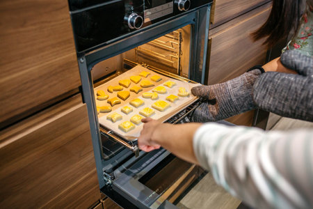 Close up of unrecognizable mom hands taking baking pan of golden cookies out of oven in kitchen while daughter pointing to biscuits with fingerの写真素材