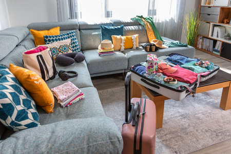 Suitcase packed with clothes and accessories for a summer vacation are placed next to a sofa in the living room, suggesting an upcoming trip. Open suitcase with colorful clothes ready for vacation.の写真素材