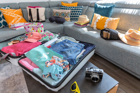 Open suitcase full of colorful summer clothes, accessories, straw hats and vintage camera lying on table, suggesting upcoming travel and wanderlustの写真素材