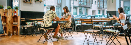 Banner of young people sitting at tables in cafe with big windows, drinking juice and working on laptop, enjoying relaxing atmosphere during their leisure timeの写真素材