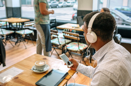 Unrecognizable young businessman wearing headphones and using smartphone while drinking cappuccino in a cafe, with digital tablet and waitress serving other customers in backgroundの写真素材