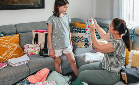Mother and daughter reviewing colorful summer clothes, folding and organizing clothing items into a suitcase, preparing for a family vacation trip, creating a sense of anticipation and excitementの写真素材
