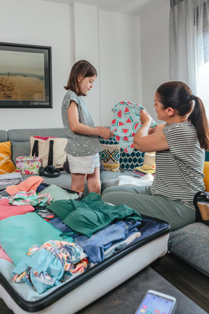 Mother and daughter are packing colorful summer clothes, folding and organizing clothing items into a suitcase, preparing for a family vacation trip, creating a sense of anticipation and excitementの写真素材