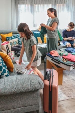 Mother and daughter folding and packing clothes into suitcases while teenage son using camera sitting on sofa in cozy living room, preparing for summer vacation trip. Family preparing bags for a trip.の写真素材