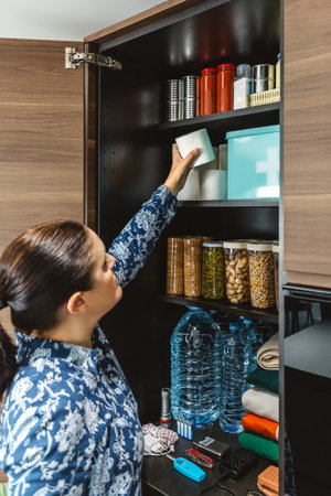 Female filling toilet paper to stocked emergency food pantry in kitchen, preparing for potential disasters. Alert, foresight and self sufficiency strategy. 72 hours survival kit concept.の写真素材