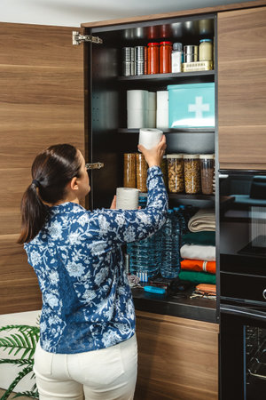 Unrecognizable female preparing 72 hours survival kit for potential disasters. Woman placing toilet paper to stocked emergency food pantry in kitchen. Alert, foresight and self sufficiency strategy.の写真素材