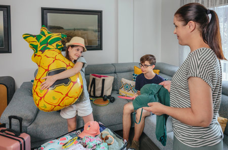 Smiling daughter hugging inflatable pineapple float while family preparing luggage for summer holidays in living room. Mother folding swimwear to suitcase trip and son playing with ping pong paddles.の写真素材