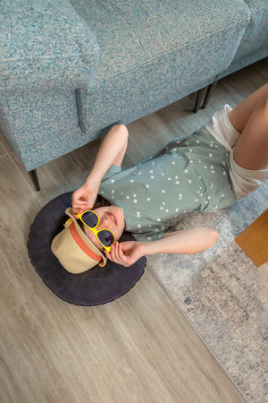 Happy young girl lying on the floor beside a sofa, wearing stylish yellow sunglasses and charming...