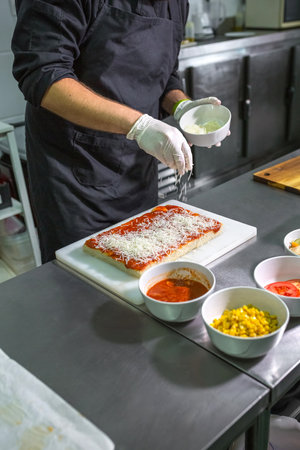 Anonymous male chef in gloves sprinkling grated cheese over a tomato sauced pizza dough on stainless steel counter, surrounded by bowls of fresh ingredients in a professional kitchenの写真素材