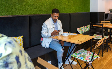 Young adult man sitting comfortably at a wooden table in coffee shop, checking his smartphone with a cup of coffee nearby, representing concepts of relaxation, digital connection, and leisure timeの写真素材