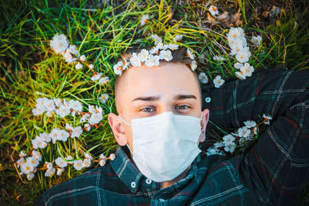 medical mask as protection against coronavirus. guy lies on grass around early spring flowers. concept of lonelinessの写真素材
