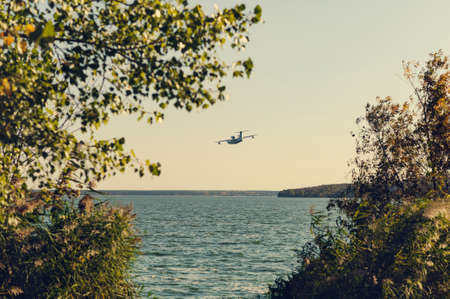 distant plane flying in the sky against the background of water and autumn plantsの写真素材