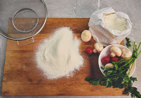 Top view of the board on which flour is poured, next to a paper bag with flour and steel sieve, a plate with tomatoes, mushrooms and greensの写真素材