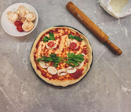 top view on a partially cooked pizza with laid out products in the form of a smile with a mustache, a number of mushrooms and kitchen accessoriesの写真素材