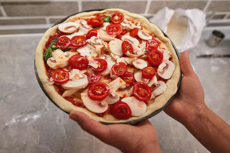 Raw pizza in the hands of a cook is ready for baking, in the background is a kitchen table with objectsの写真素材