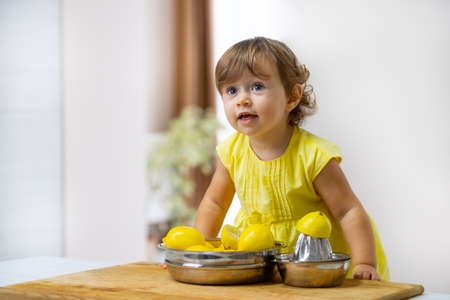 Little girl in a yellow dress prepares lemonade using a citrus press while sitting in the kitchenの写真素材