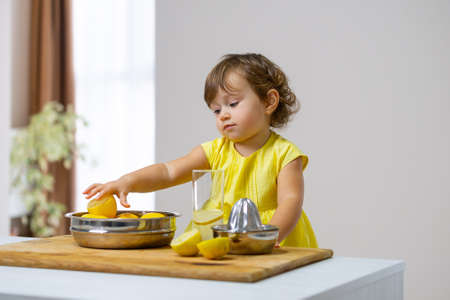 Little girl in a yellow dress prepares lemonade using a citrus press while sitting in the kitchenの写真素材