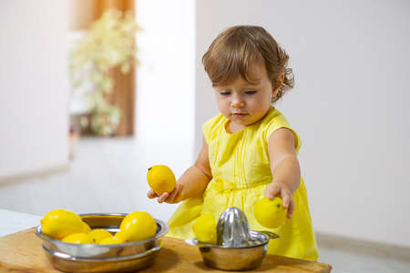 Little girl in a yellow dress prepares lemonade using a citrus press while sitting in the kitchenの写真素材