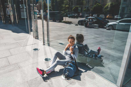 Young woman sits with her back elbows against a glass wall and touches headphones with her handsの写真素材